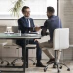 Smiling multiracial young businessmen handshake greeting get acquainted at boardroom briefing, excited diverse male business partners shake hands make agreement after successful office negotiations