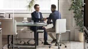 Smiling multiracial young businessmen handshake greeting get acquainted at boardroom briefing, excited diverse male business partners shake hands make agreement after successful office negotiations