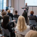 Business conference presenter speaking to attentive audience in modern meeting room, networking, learning, professional seminar atmosphere.