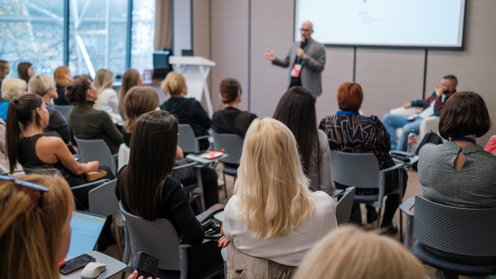 Business conference presenter speaking to attentive audience in modern meeting room, networking, learning, professional seminar atmosphere.