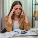 Young woman at home table rests forehead in hands staring at overdue bills laptop and calculator feeling anxious. Caucasian girl battles worry trying to figure payment plan before deadlines arrive.