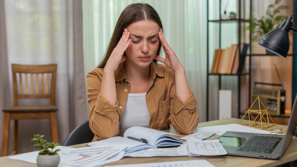 Young woman at home table rests forehead in hands staring at overdue bills laptop and calculator feeling anxious. Caucasian girl battles worry trying to figure payment plan before deadlines arrive.