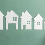 A human hand points to a small paper house cutout in a row of varying home sizes on a green background
